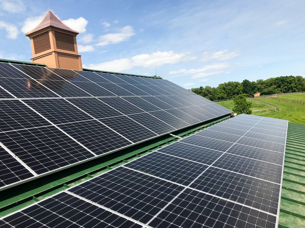 Rooftop view of over five dozen solar panels on farm building with view of fenced tree-lined grassy land - Residential Solar system installation by top Solar Installer In Willow Street PA